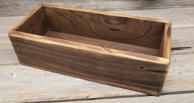 May include: A rectangular wooden box with a dark brown finish. The box is open at the top and appears to be handmade. The wood grain is visible, adding to the rustic aesthetic. The box is likely used for storage or display.