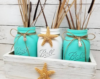 May include: Three turquoise blue mason jars with white lids and twine tied around the top. The jars are filled with dried grasses and a starfish. The jars are sitting in a white wooden box with a starfish on the front. The box is sitting on a white wooden surface.