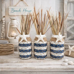 May include: Three decorative mason jars with blue and white stripes, adorned with twine, netting, and starfish. Each jar holds dried wheat stalks. The jars have the words "Beach House" on them.