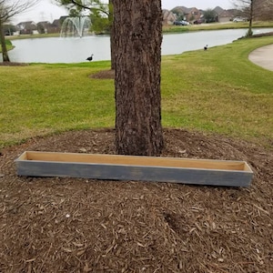May include: A rectangular wooden planter box with a gray exterior and natural wood interior sits at the base of a tree. The planter is long and narrow, designed for outdoor use. The background includes a lake and houses.
