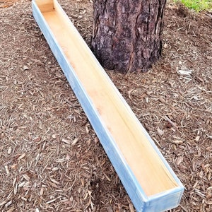 May include: A long, rectangular wooden planter box with a light blue painted exterior and natural wood interior. The planter is empty and sits on a bed of wood chips near a tree trunk. The box is designed for outdoor use.
