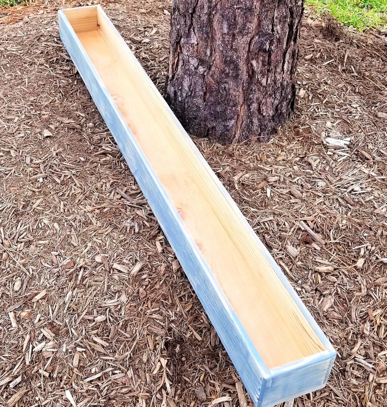 May include: A long, rectangular wooden planter box with a light blue painted exterior and natural wood interior. The planter is empty and sits on a bed of wood chips near a tree trunk. The box is designed for outdoor use.