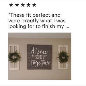 Pode incluir: Uma placa de madeira cinza com letras brancas que diz "Home is wherever we're together". A placa est&aacute; pendurada em uma parede com duas molduras de janelas de madeira branca de cada lado. Cada moldura de janela tem uma coroa verde.