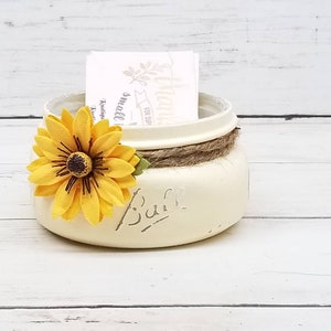 May include: Cream-colored mason jar with a yellow sunflower attached. The jar has a twine band around the middle and the word "Ball" is printed on the front. A small card is inside the jar.