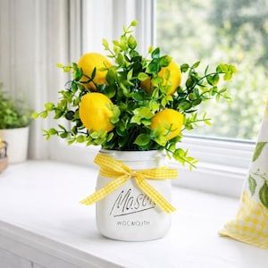 May include: A white Mason jar with a yellow ribbon bow, filled with faux lemons and green foliage. The jar is on a white windowsill, with a tiered tray of lemons and a lemon-themed curtain in the background. The jar reads "Mason Wide Mouth."