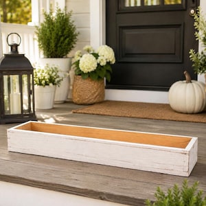 May include: A long, rectangular wooden planter box with a distressed white painted exterior finish and a natural light brown wood interior. The container features straight sides and a flat base, designed for holding plants or decorative items.