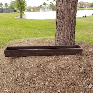 May include: A long, rectangular wooden planter box with a dark brown stain. The planter is sitting on a bed of brown wood chips.