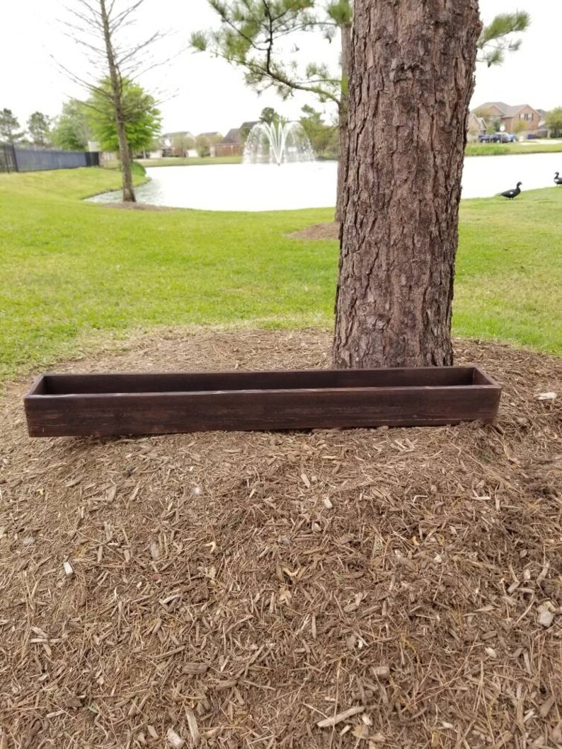 May include: A long, rectangular wooden planter box with a dark brown stain. The planter is sitting on a bed of brown wood chips.