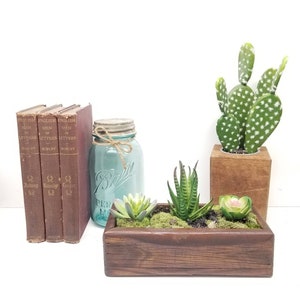 May include: A rustic home decor arrangement featuring three vintage books, a turquoise mason jar, and a wooden planter box with succulents. A green cactus in a wooden block adds a natural touch.