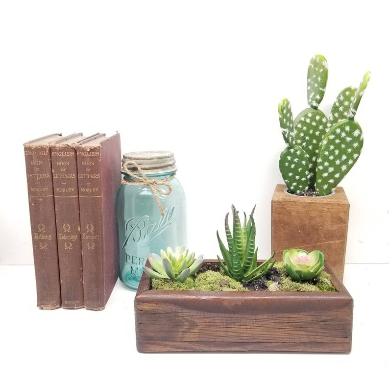May include: A rustic home decor arrangement featuring three vintage books, a turquoise mason jar, and a wooden planter box with succulents. A green cactus in a wooden block adds a natural touch.