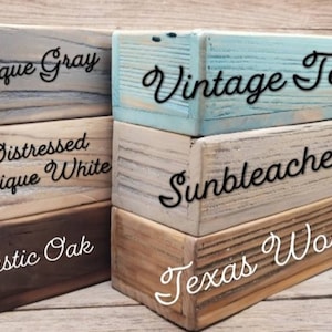 May include: Stacked wooden blocks painted in various colors and distressed finishes. The blocks are labeled with the paint colors: Antique Gray, Distressed Antique White, Rustic Oak, Vintage Teal, Sunbleached, and Texas Wood.