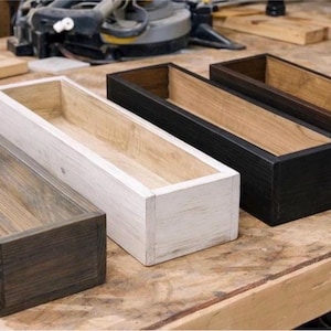 May include: Four rectangular wooden planters in varying colors: dark gray, white, black, and brown. Each planter has a natural wood interior and a stained exterior. The planters are arranged on a wooden workbench.