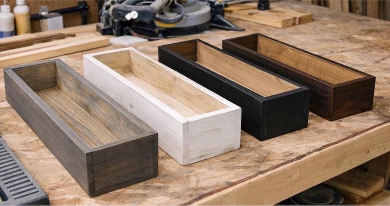 May include: Four rectangular wooden planters in varying colors: dark gray, white, black, and brown. Each planter has a natural wood interior and a stained exterior. The planters are arranged on a wooden workbench.