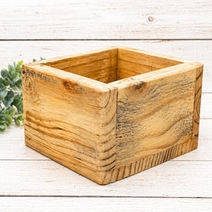 May include: A square wooden box with a natural wood finish. The box is constructed from planks of wood, showcasing the grain and knots. The box is empty and has a rustic, handcrafted appearance.