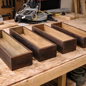 May include: Four rectangular wooden boxes with a dark brown exterior and a lighter wood interior sit on a wooden workbench. The boxes are empty and appear to be handmade. The workbench is also made of wood.