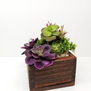 May include: A small wooden planter box filled with artificial succulents. The succulents are various shades of green and purple. The box is a dark brown color and has a square shape.