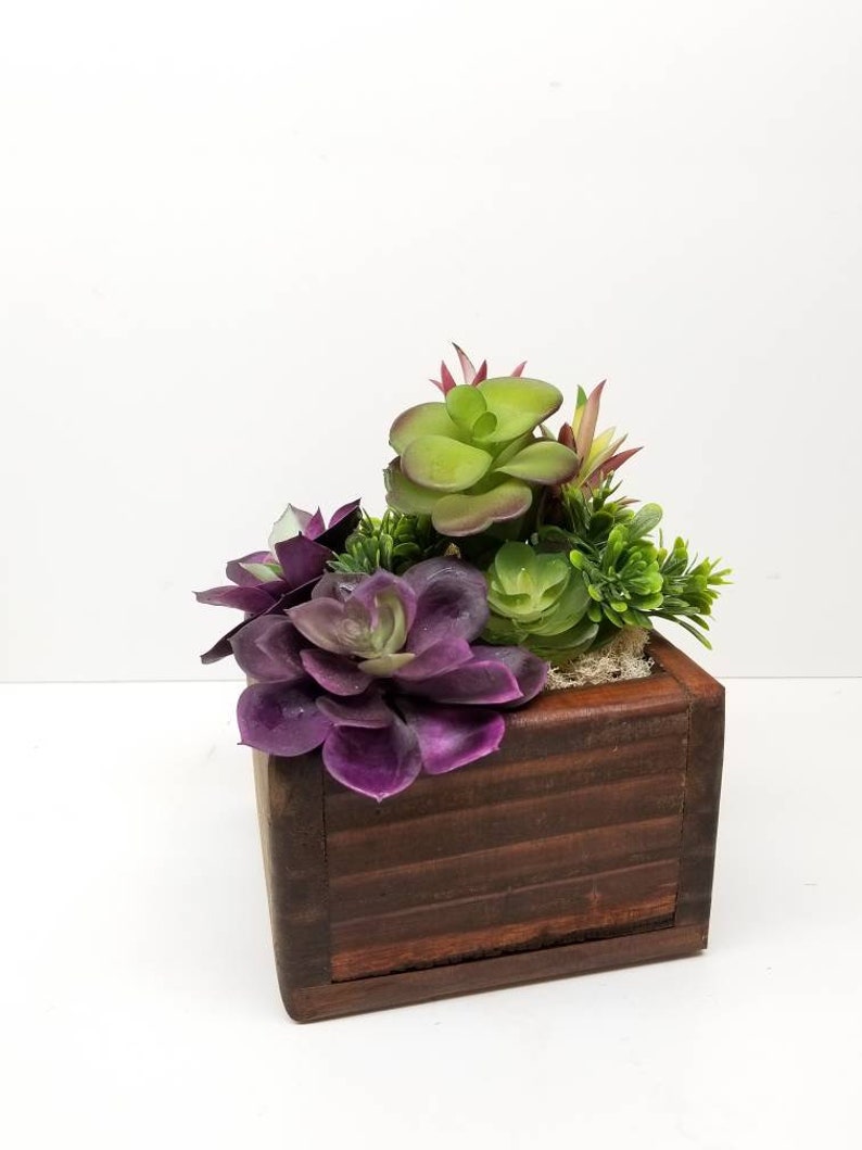 May include: A small wooden planter box filled with artificial succulents. The succulents are various shades of green and purple. The box is a dark brown color and has a square shape.