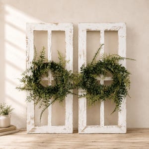 May include: Two rectangular white wooden window frames with a distressed finish, each featuring a green artificial fern wreath attached to the center. The frames have a vertical grid design and are positioned side by side against a neutral wall.