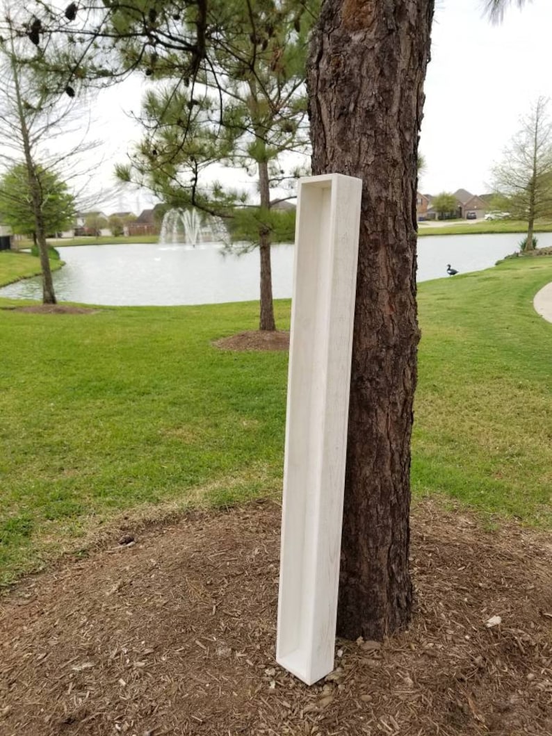 May include: A long, white wooden planter box with a rustic finish. The planter is leaning against a tree trunk in a park setting.