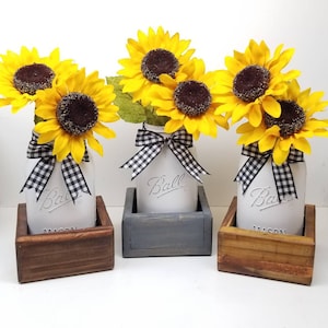May include: Three mason jars with yellow sunflowers and black and white checkered bows. The jars are in wooden boxes of varying colors. The jars have the word "Ball" embossed on them.
