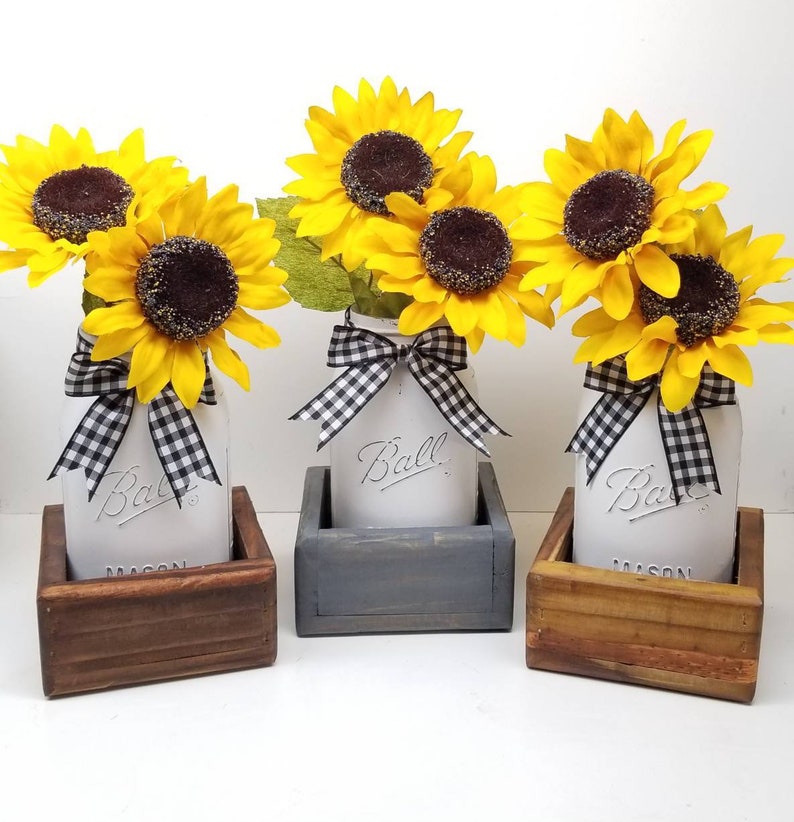 May include: Three mason jars with yellow sunflowers and black and white checkered bows. The jars are in wooden boxes of varying colors. The jars have the word "Ball" embossed on them.