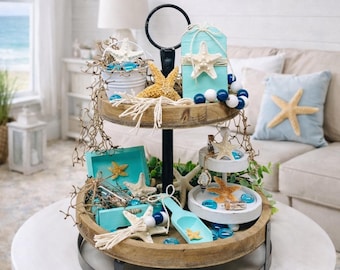 May include: A two-tiered wooden tray with a coastal theme. The tray is decorated with starfish, blue glass beads, and small turquoise boxes. A starfish-adorned pillow and framed starfish art are in the background, creating a beachy aesthetic.