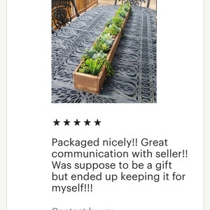 May include: A long, rectangular wooden planter box filled with a variety of artificial succulents and greenery. The box is stained a warm brown and sits on a decorative metal table. The arrangement includes various shades of green and gray.
