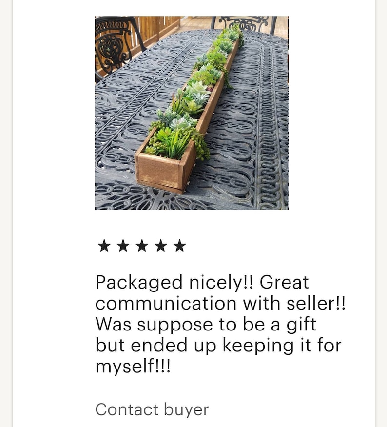 May include: A long, rectangular wooden planter box filled with a variety of artificial succulents and greenery. The box is stained a warm brown and sits on a decorative metal table. The arrangement includes various shades of green and gray.