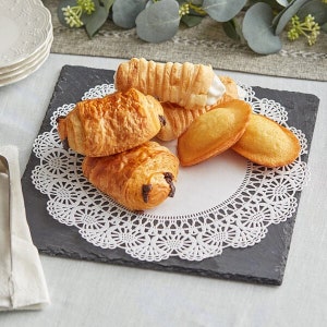 May include: A white doily sits on a black slate plate with a variety of pastries, including croissants, madeleines, and chocolate-filled pastries.
