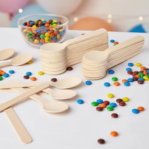 May include: A clear glass bowl filled with colorful candies sits next to stacks of small wooden spoons. Scattered around are more candies in various colors. The spoons are light brown and appear to be disposable.