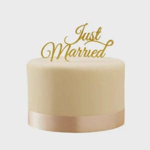 May include: A round, cream-colored cake with a gold ribbon around the base. A gold glitter "Just Married" cake topper is placed on top. The cake is a simple design, perfect for a wedding or anniversary celebration.