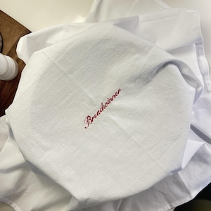 May include: A white kitchen towel draped over a round object, featuring the word Breadwinner embroidered in a red cursive script font. The fabric has a visible woven texture and is folded to create soft, natural creases across the surface.