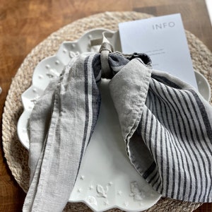 May include: A white ceramic plate with a scalloped edge and a floral design. A gray and white striped linen napkin is tied around the plate with a brown cord. The plate is on a woven straw placemat.