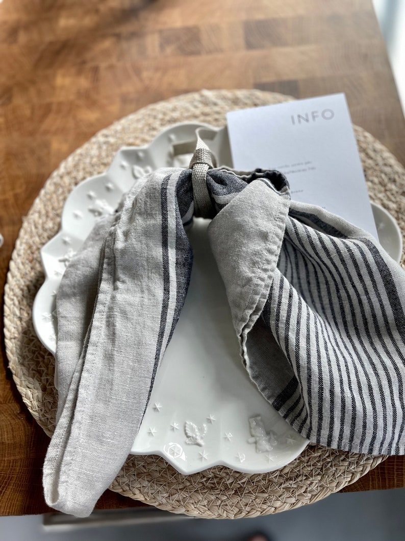 May include: A white ceramic plate with a scalloped edge and a floral design. A gray and white striped linen napkin is tied around the plate with a brown cord. The plate is on a woven straw placemat.