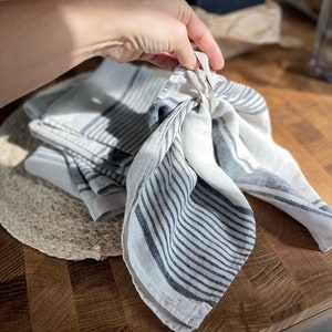 May include: A set of three white linen kitchen towels with black stripes. The towels are folded and tied together with a white string.