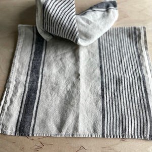 May include: A white linen kitchen towel with black stripes. The towel is folded in half and is lying on a wooden surface.