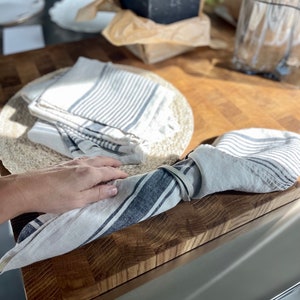 May include: A set of two white linen kitchen towels with blue stripes. The towels are folded and tied together with a white linen ribbon. The towels are on a wooden cutting board.