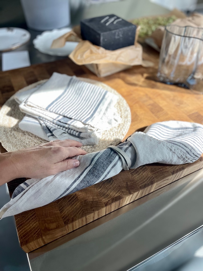 May include: A set of two white linen kitchen towels with blue stripes. The towels are folded and tied together with a white linen ribbon. The towels are on a wooden cutting board.
