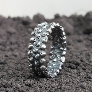 May include: A silver ring featuring a repeating pattern of tiny skulls. The ring has a textured, detailed surface and a dark, oxidized finish. The ring is displayed on a dark, textured surface.