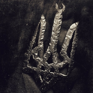 May include: A sepia-toned image of a textured metal trident, the national symbol of Ukraine. The trident has three prongs and a curved top, set against a dark, textured background. The metal appears to have a rough, hammered finish.