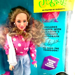 May include: A pink and white polka dot shirt, blue denim shorts, and a white tote bag are worn by a doll with brown hair and a pink headband. The doll is holding a purple and white yearbook with the text "Bayside High School Yearbook". The doll is a replica of the character Jessie from the television show Saved by the Bell.