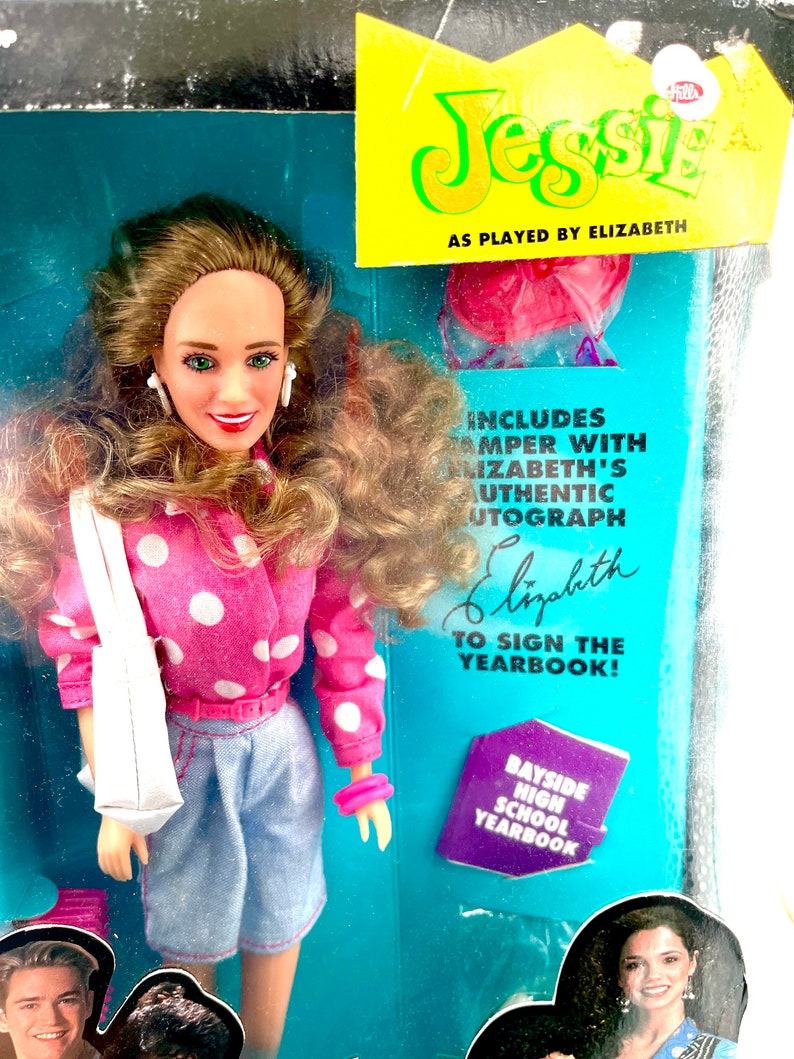 May include: A pink and white polka dot shirt, blue denim shorts, and a white tote bag are worn by a doll with brown hair and a pink headband. The doll is holding a purple and white yearbook with the text "Bayside High School Yearbook". The doll is a replica of the character Jessie from the television show Saved by the Bell.