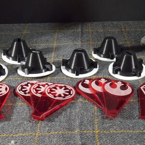 May include: A selection of black and white game pieces, alongside red translucent tokens with white symbols. The items are displayed on a grey patterned surface, possibly for a tabletop game or hobby.