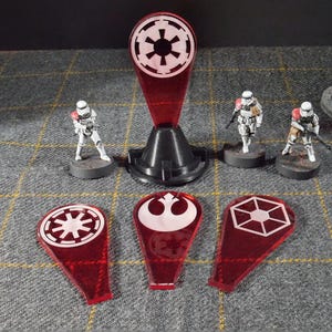 May include: A tabletop game setup with miniature Stormtrooper figures and red acrylic tokens with white symbols. A black stand holds a larger red token. The scene is set on a grey, chequered surface with miniature castle structures in the background.
