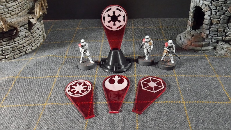 May include: A tabletop game setup with miniature Stormtrooper figures and red acrylic tokens with white symbols. A black stand holds a larger red token. The scene is set on a grey, chequered surface with miniature castle structures in the background.