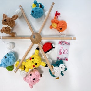 May include: A wooden mobile with crocheted Pokemon characters, including Pikachu, Bulbasaur, Charmander, Squirtle, and a brown character. The mobile has white balls and a pink card with the text "Thank You! Zummy".