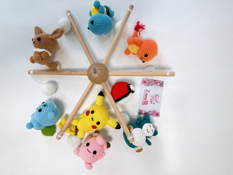 May include: A wooden mobile with crocheted Pokemon characters, including Pikachu, Bulbasaur, Charmander, Squirtle, and a brown character. The mobile has white balls and a pink card with the text "Thank You! Zummy".
