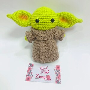 May include: A green crocheted plush toy with large ears and brown crocheted clothing. The toy is sitting on a white surface with a thank you card that says "Thank you" and "Z inny" with a yarn ball icon.