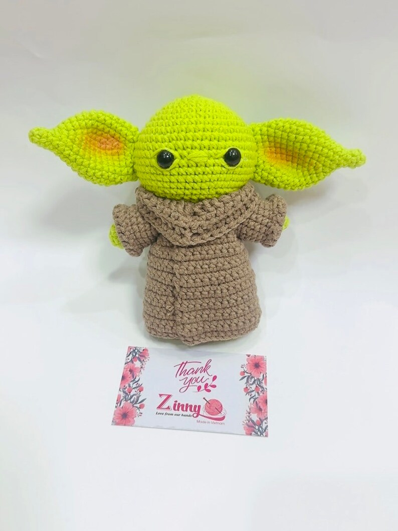 May include: A green crocheted plush toy with large ears and brown crocheted clothing. The toy is sitting on a white surface with a thank you card that says "Thank you" and "Z inny" with a yarn ball icon.