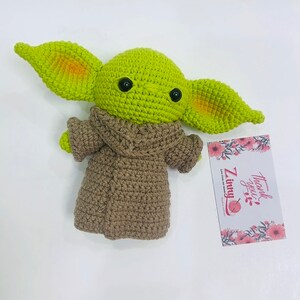 May include: Green crocheted plush toy with large ears and brown crocheted clothing. The toy is sitting on a white surface with a thank you card.